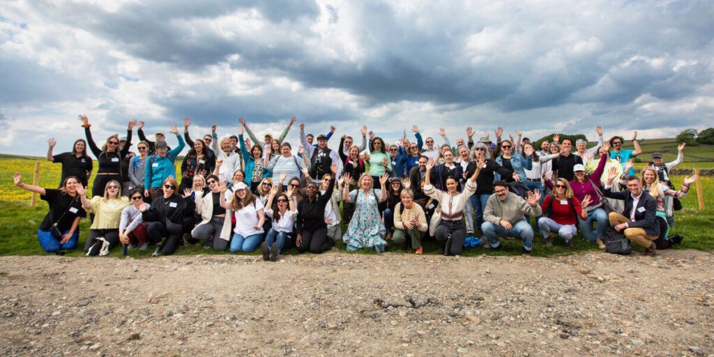 A large group of funders are posing for a photo and cheering in a field with a bright cloudy sky.