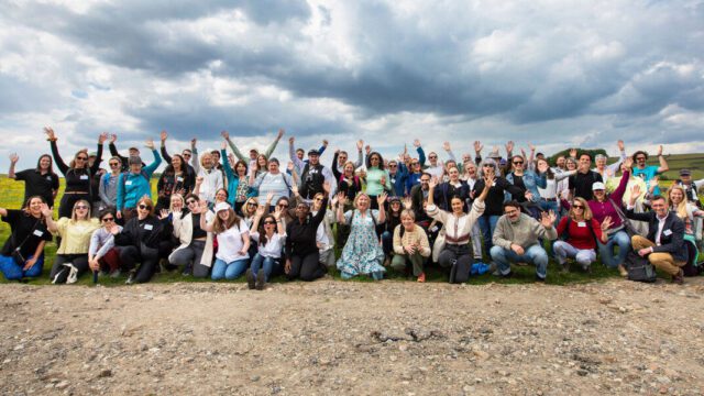 A large group of funders are posing for a photo and cheering in a field with a bright cloudy sky.
