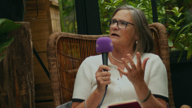 An older white woman with a short grey bob, Elizabeth, is speaking into a purple microphone with the EFN logo on whilst sitting in a wicker chair in a greenhouse.