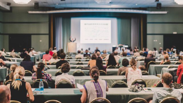 A large group of people are sitting at tables watching a presentation on the stage.