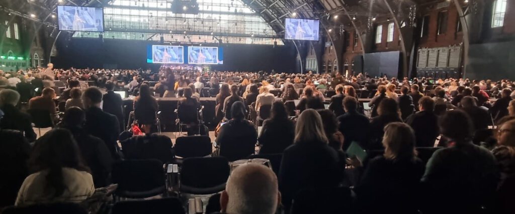 A large group of people are sitting in a big conference room watching a speaker and presentation on a stage