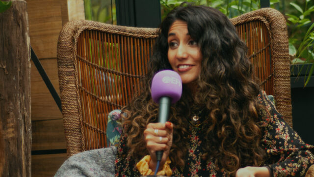 A brown woman with long, curly black hair, Aruna, is speaking into a purple microphone with the EFN logo on whilst sitting in a wicker chair in a greenhouse