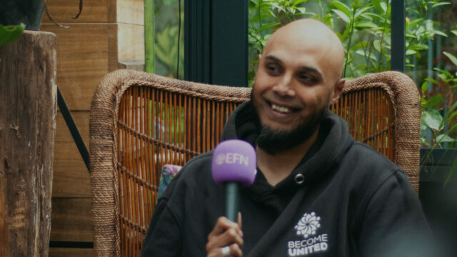 A bald, brown man with a black beard and a black hoodie, is speaking into a purple microphone with the EFN logo on whilst sitting in a wicker chair in a greenhouse