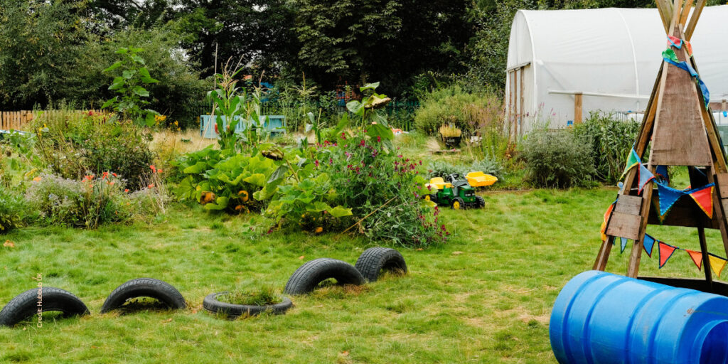 A community garden with a lawn, black tyre path markets, a brown teepee and a white fabric greenhouse tunnel.