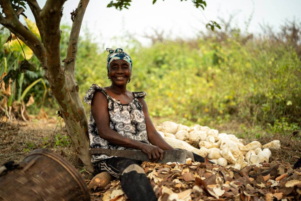 Christine, a black woman wearing a pattern top and a colourful hair wrap, is sitting down, smiling to camera whilst she peels cassava.