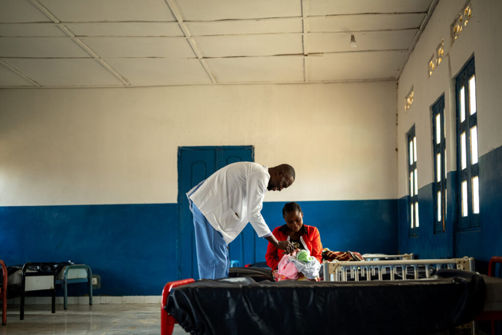 Bakumba, a doctor wearing scrubs and a white lab coat, is speaking and checking a newborn baby, wrapped in a pink blanket, and mother, Augustine, sitting and smiling, in a plain hospital ward in the DRC.