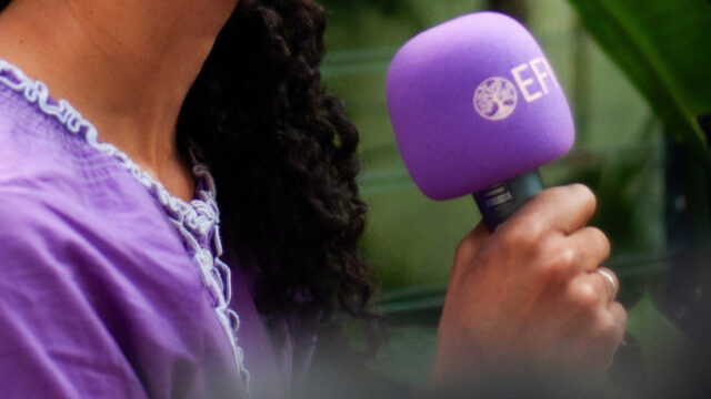 A close up of a brown hand and arm holding a microphone with a purple cover and the EFN logo on it.