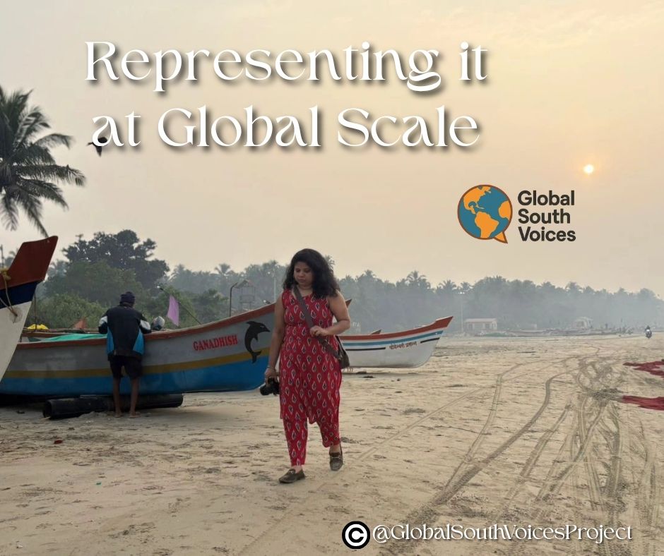 In Malvan on India’s Konkan coast, an Indian woman, Gayatri, walks along a beach in a red punjabi suit. There are fishermen and boats behind her.
