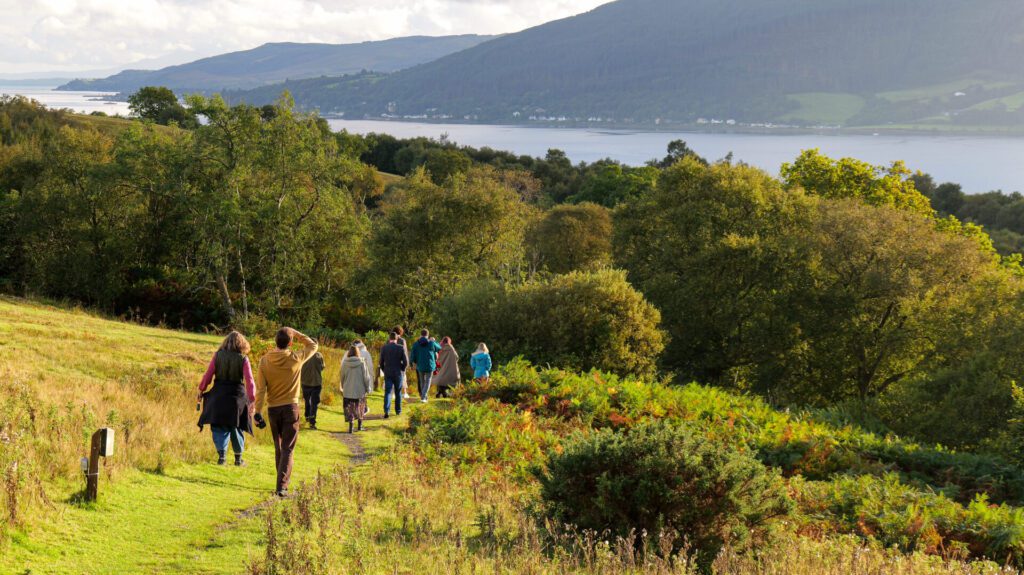 A group of funders walk down a hill in the golden sunshine with a loch and forest in the background.