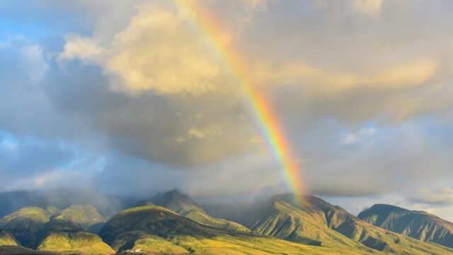 A rainbow in a grey sky appears over a landscape of large rolling green hills and mountains