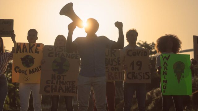 A group of protestors of different backgrounds and ages are standing in a line holding protest signs and a bullhorn. They are shaded by the sun behind them.
