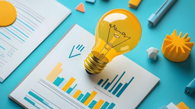A graphical yellow lightbulb sits on top of a background of reports with graphs and stationary on a blue table.