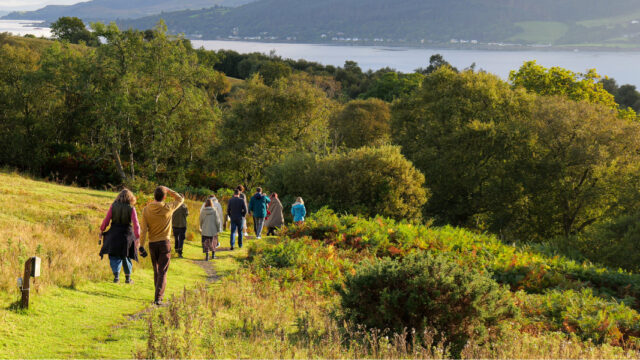 A group of men and women walk down a green and forested hill with a Scottish loch in the distance