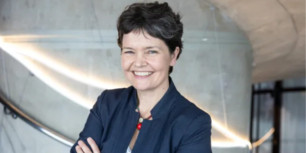 A portrait of Kate Raworth, a white older woman with short brown hair. She is smiling to camera with her arms crossed, a blue top in front of a plain wall background