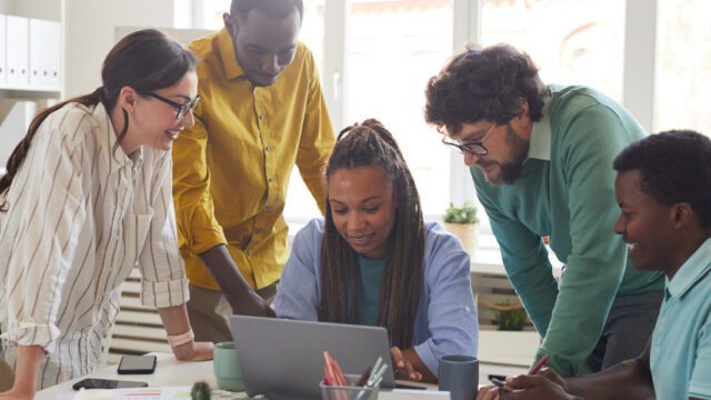 A group of people are discussing work around a laptop. There is a black woman sitting by the laptop and her colleagues are one white woman, two black men and one brown man, all in casual office clothes.