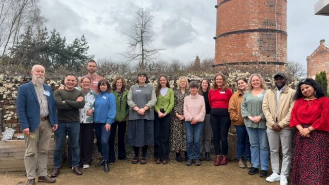 A mixed group of about 30 men and women are smiling to the camera in outdoor area with a large brick chimney above them.