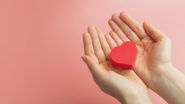 A white person's hands holding out a paper red heart